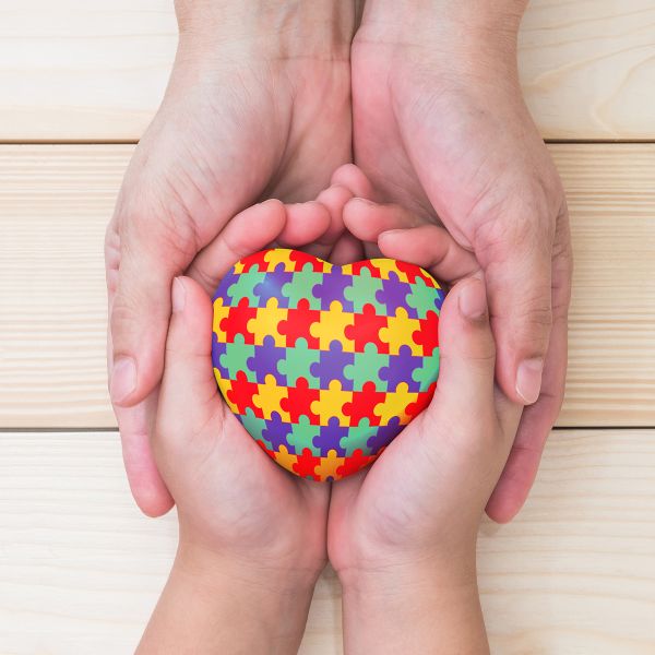 An adult pair of hands holding a child's hands, which are holding a multi-coloured heart